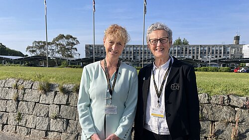Cllrs Hillary Frank and Sarah Preece outside, with a stone wall, a grassy field with flagpoles and a building behind them