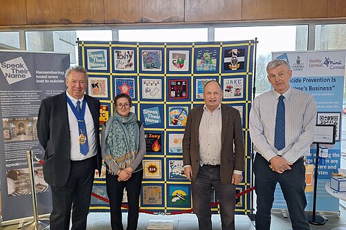 Four people, including Cornwall Liberal Democrat councillors Rob Nolan and Jim Candy, stand in front of information boards. The leftmost board gives information about Speak Their Name, an organisation remembering loved ones lost to suicide. The centre board displays pictures created in support of Speak Their Name, the rightmost board shows Cornwall Council's initiatives for suicide prevention.