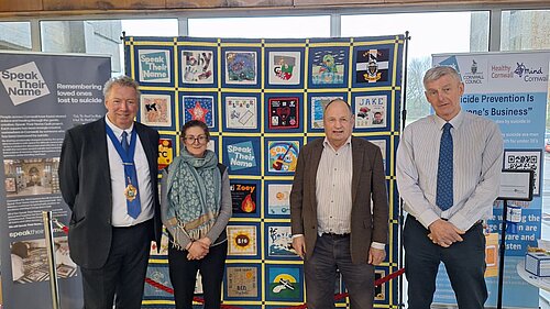 Four people, including Cornwall Liberal Democrat councillors Rob Nolan and Jim Candy, stand in front of information boards. The leftmost board gives information about Speak Their Name, an organisation remembering loved ones lost to suicide. The centre board displays pictures created in support of Speak Their Name, the rightmost board shows Cornwall Council's initiatives for suicide prevention.