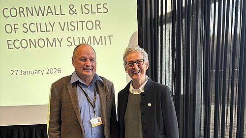 Cornwall councillors Jim Candy and Sarah Preece stand in front of a slide projection. The slide text reads: Cornwall & Isles Of Scilly Visitor Economy Summit. 