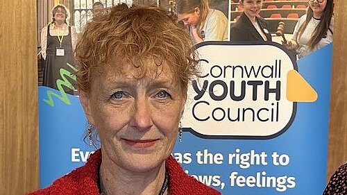 A woman with short red hair stands in front of a poster. Text of the poster reads Cornwall Youth Council.