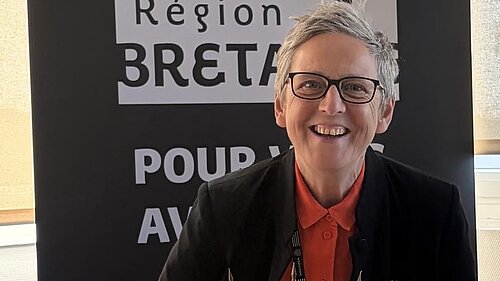 Cllr Sarah Preece in front of a banner saying 'Région Bretagne'. The tops of flags representing the Celtic nations, including Cornwall and Brittany, are just visible at the bottom