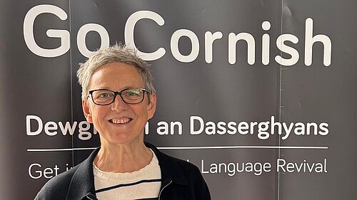 Cllr Sarah Preece in front of a banner saying 'Go Cornish', and two lines of partially obscured text; the first in Cornish: 'Dewg... an Dasserghyans', the second: 'Get... Language Revival'