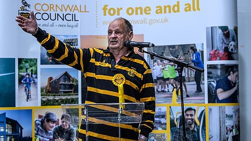 Cornwall Councillor Jim Candy, dressed in Cornish colours, gestures by a microphone. In the background is a collage of pictures, topped by Cornwall Council for one and all.