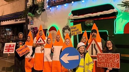 People dressed as traffic cones stand outside a pub. They are holding a variety of placards designed to resemble road signs.